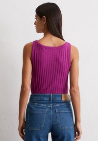 Dopasowany, bezrękawnik z prążkowanej tkaniny w kolorze magenta, połączony z wysokim stanem niebieskich dżinsów. Widoczne szwy i teksturowane akcenty.
