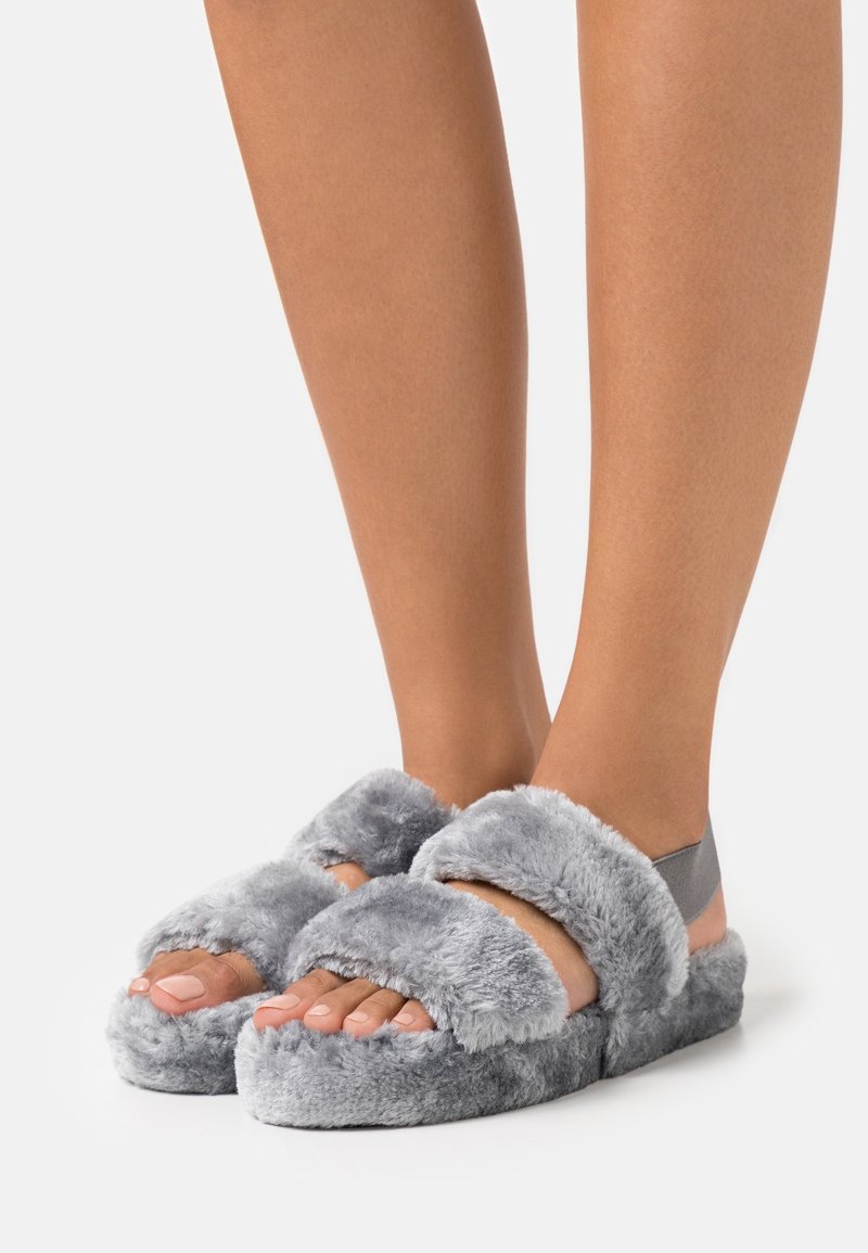 Pieds portant des chaussons ouverts gris duveteux avec deux larges brides et une bride élastique grise au talon sur fond blanc.