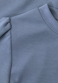 Vaaleansininen pitkähihaine paita, jossa on pyöreä kaula-aukko ja hieman poimutettu puffiyksityiskohta olkapäässä.