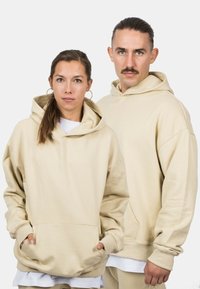 Beige hoodies with a relaxed fit and large hoods, featuring kangaroo pockets and ribbed cuffs, worn with white shirts underneath.