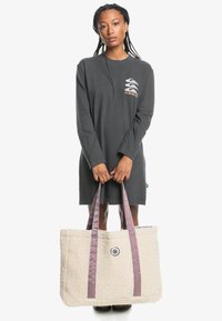 Große Creme-Totetasche aus einem flauschigen Textil, mit zwei lila Stoffgriffen und einem runden Knopfdetail auf der Vorderseite.