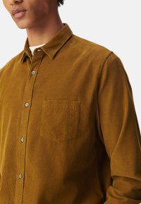 Bruine corduroy overhemd met een button-down kraag, voorzien van een borstzak en een gestructureerd oppervlak. Knopen zijn zichtbaar en complementair van kleur.