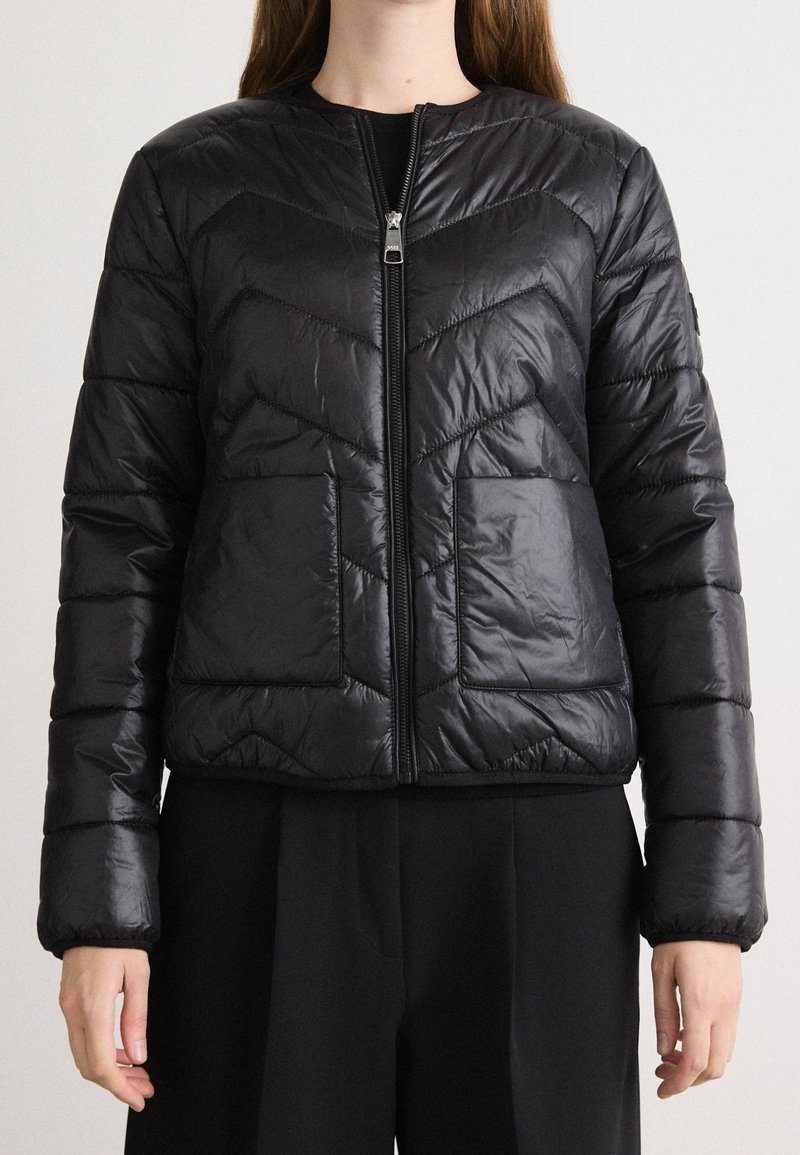 Femme portant une veste noire matelassée zippée avec manches longues et deux poches avant, associée à un pantalon noir.