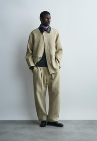 Mand stående med hænder i lommerne iført beige oversize jakke og bukser med sort krave og sorte sko foran en ensfarvet væg.