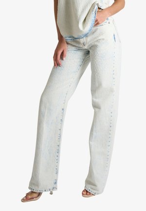 Donna che indossa jeans dritti lavati all'acido azzurro chiaro e sandali con tacco trasparente, con una mano in tasca e l'altra che riposa sulla coscia.