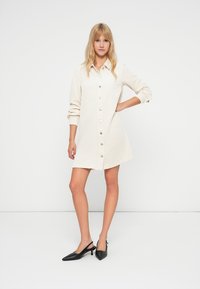 Crèmekleurige corduroy jurk met lange mouwen, knopen aan de voorkant en een relaxte pasvorm, gecombineerd met zwart, puntige pumps.