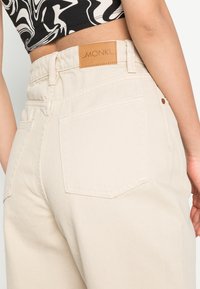 Calças de tecido bege com cintura alta, apresentando bolsos cosidos e uma etiqueta de couro gravada com "MONKI" na parte de trás da cintura.