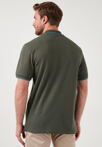 Temnozeleni polo majica s kratkimi rokavi, ki vsebuje teksturirano tkanino in turkizno poudaritev okoli ovratnika in robov rokavov.