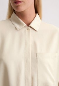 Helles beige Hemd mit Kragen, Knopfverschluss, einer Brusttasche, glattem Stoff mit dezentem Glanz, präsentiert sich mit klaren Linien und minimalistischem Design.