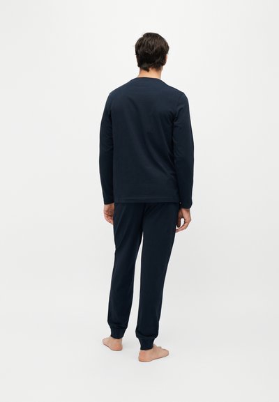Homme debout pieds nus, de dos, portant une chemise à manches longues foncée et un pantalon assorti sur un fond clair uni.