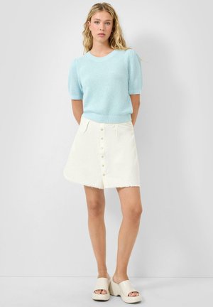 Kobieta stoi, ubrana w jasnoniebieski, teksturowany sweter z krótkim rękawem, białą spódnicę na guziki i beżowe sandały na platformie na białym tle.