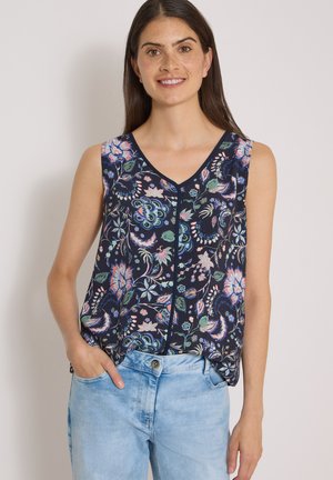 Femme portant un haut bleu marine sans manches avec un motif floral multicolore et un jean bleu clair, une main dans la poche.
