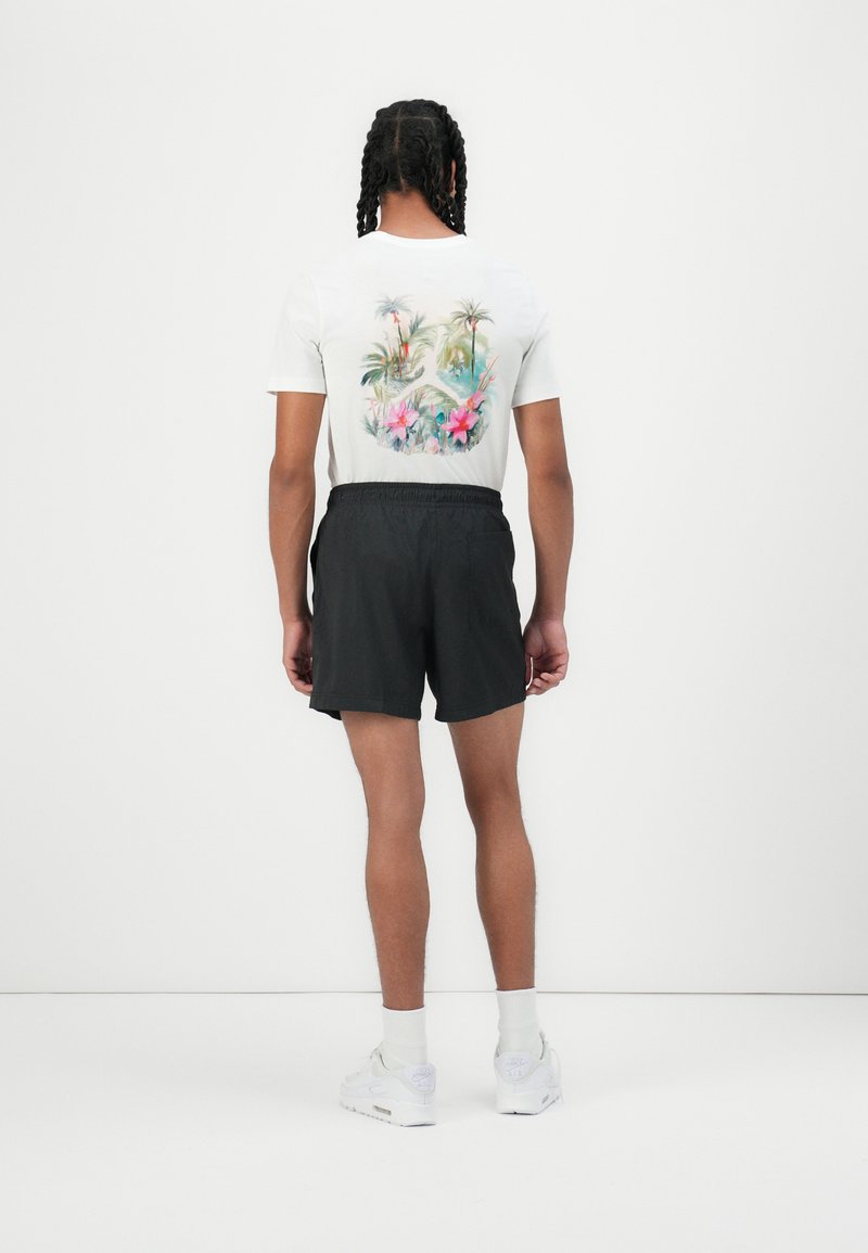 T-shirt blanche avec un imprimé tropical coloré au dos, associée à un short noir et des baskets de sport blanches avec des chaussettes grises claires.