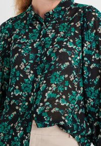Camicia con bottoni in tessuto nero trasparente con un motivo floreale verde e crema. Vestibilità ampia con colletto e maniche con polsino.