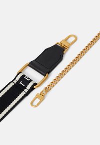 Correa de tela negra y crema con herrajes dorados y un accesorio de cadena texturizada. Presenta dos extremos con clip y un detalle de logo en la correa.
