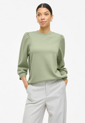 Light green long-sleeve top with puffed shoulder seams, round neckline, and smooth texture. Paired with light gray tailored trousers.
