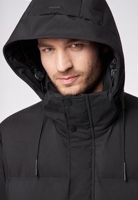 Giacca nera isolata con collo alto e cappuccio regolabile. Presenta bottoni a pressione e una texture liscia. Design semplice senza fantasie.