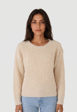 Jonge vrouw met lang bruin haar, draagend een beige gebreide trui met knopen op de schouder en lichtblauwe jeans, staand tegen een effen achtergrond.