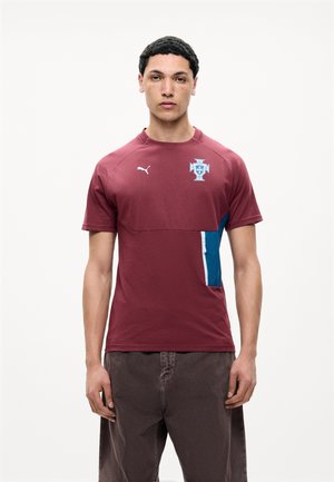 Junger Mann mit kurzen lockigen Haaren, der ein bordeauxfarbenes Puma-Sportshirt mit blau-weißem Abzeichen und dunkle Hose trägt, steht vor weißem Hintergrund.