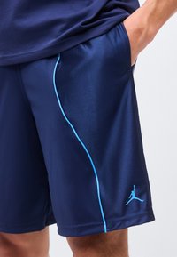 Shorts de sport bleu marine avec un liseré bleu clair et un logo Jumpman sur la jambe, portés avec une main dans la poche.