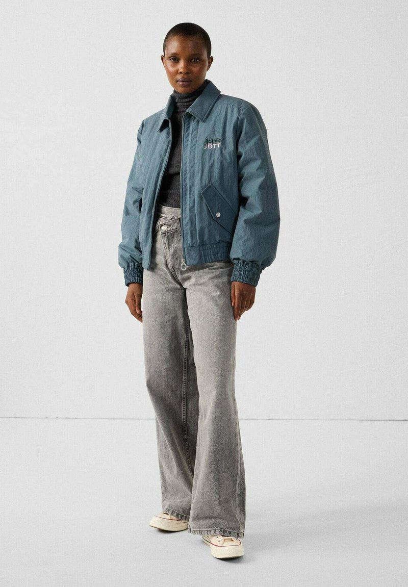Blauwe bomberjack met elastische manchetten, gecombineerd met lichtgrijze flared jeans. De jas heeft een borstzak en minimale branding.