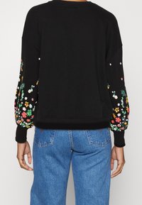 Sweat-shirt noir avec broderie florale colorée sur les manches, col rond et poignets côtelés. Le dos présente une texture lisse et une coupe décontractée.