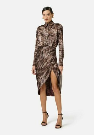 Donna che indossa una camicia a maniche lunghe marrone con motivi e una gonna coordinata con spacco alto, tiene una pochette e indossa tacchi neri con cinturino alla caviglia.