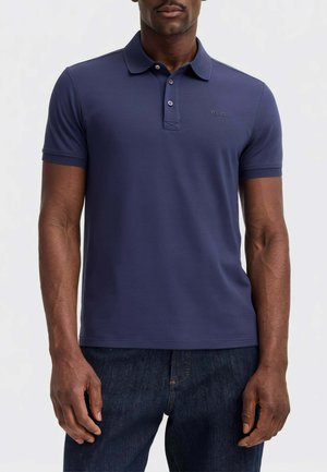 Modello maschile che indossa una polo blu navy a maniche corte e jeans blu scuro, in piedi davanti a uno sfondo chiaro e uniforme.