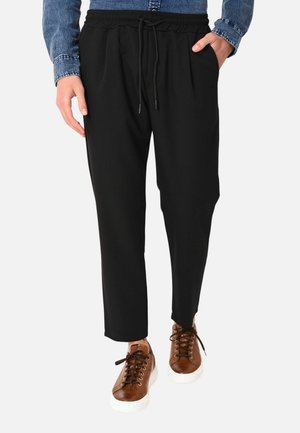 Persona che indossa pantaloni neri con coulisse, scarpe da ginnastica marroni con lacci e suole bianche, e una camicia di jeans blu con una mano in tasca.