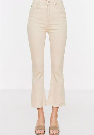 Beige high-waisted cropped flare jeans gedragen met taupekleurige open-toe sandalen met hakken op een witte achtergrond.