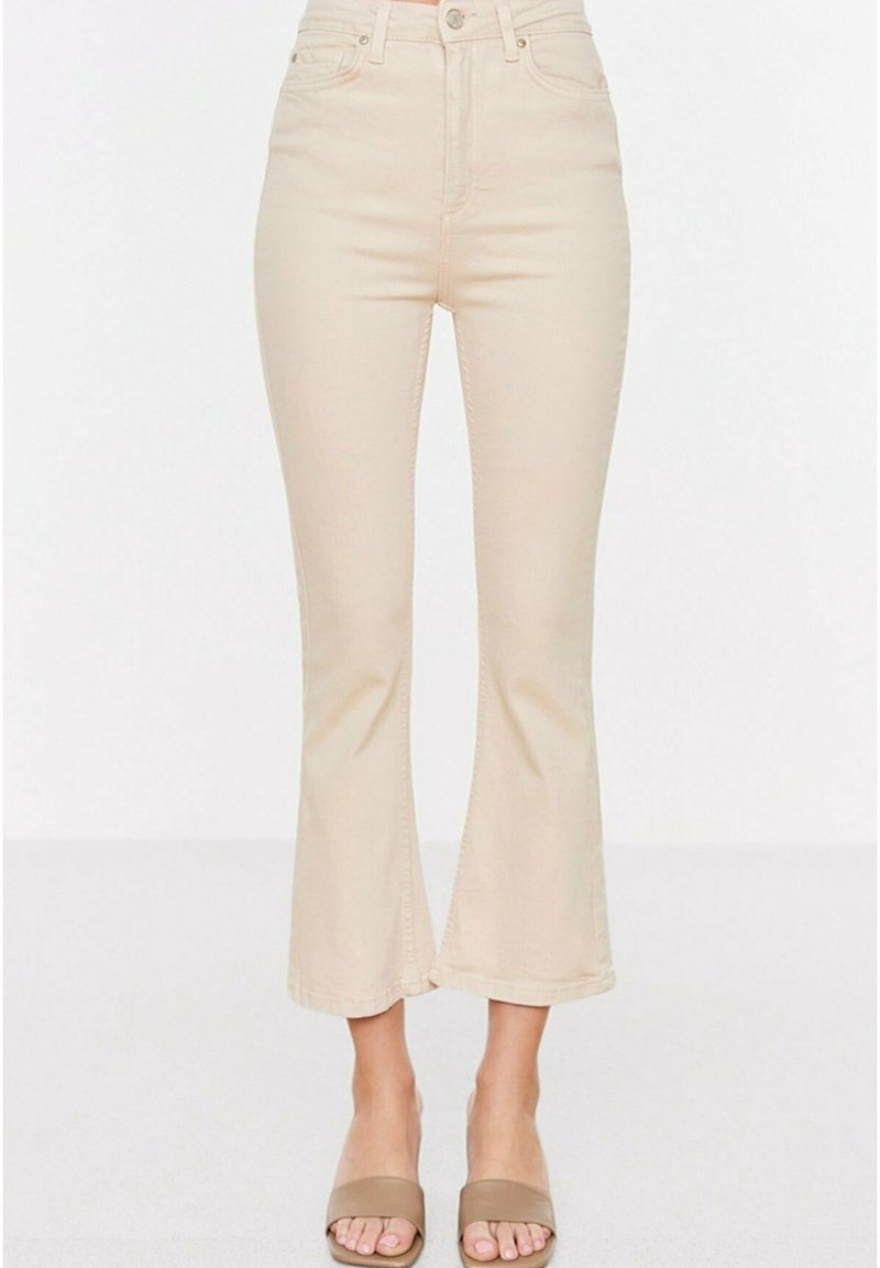 Jean beige taille haute coupé flare, porté avec des sandales à talons ouvertes taupe sur fond blanc.