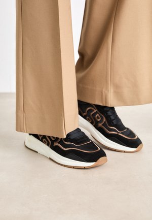 Schwarze und braune Sneaker mit weißen Sohlen, getragen zu beigen Hosen mit weitem Bein auf einem hellen Boden.