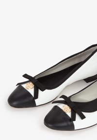 Coppia di ballerine da donna bianche e nere con punta nera, fiocchi neri e decorazioni circolari dorate sulla parte anteriore.