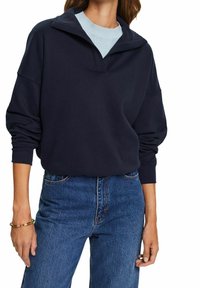 Suéter holgado de color azul marino con cuello de medio cierre y detalles acanalados; combinado con jeans de mezclilla de tiro alto. Textura suave, ajuste relajado.