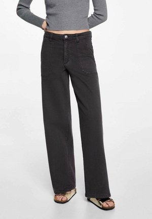 Kvinde iført sorte, højtaljede, vidde-ben jeans, grå ribstrikket langærmet cropped top og gyldne åbne tåsandaler, stående med hænderne bag ryggen.