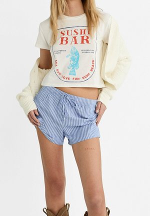 Jeune femme portant un t-shirt crop crème avec le texte "Sushi Bar", un short rayé bleu clair et un cardigan crème drapé sur ses bras.