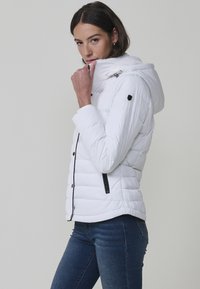 Chaqueta acolchada blanca con capucha con cremallera, detalles de hardware negro y acolchado horizontal. Presenta un diseño ajustado y bolsillos laterales con cremallera.