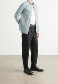 Henkilö, joka on pukeutunut vaaleansiniseen nappipaitaan valkoisen topin päälle, mustiin housuihin vyöllä ja mustiin nauhakengiin, seisoo harmaalla lattialla.