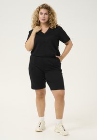 Romper nero a maniche corte con scollo a V e vita elastica, caratterizzato da una vestibilità rilassata. Indossato con sneakers bianche sportive. Design minimalista.