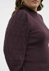 Donna che indossa un maglione lavorato a maglia viola con maniche a sbuffo a motivo testurizzato e orecchini a cerchio dorati, mostrata dalla zona del collo fino a metà torso.