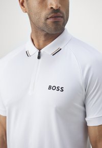 Man wearing a white polo shirt with a zip collar and "BOSS" logo on the chest, shown from shoulders to chin against a plain background.