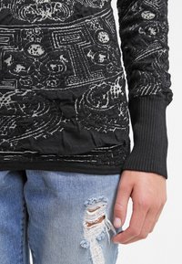 Haut noir à manches longues avec des motifs texturés complexes, associé à un jean bleu clair déchiré avec une déchirure effilochée sur la cuisse.