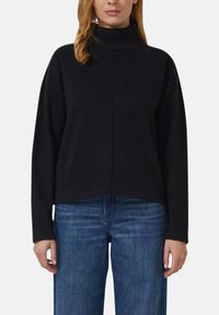 Schwarzer Pullover mit hohem, geripptem Kragen, weiten Ärmeln und kurzem Schnitt. Weicher Stoff mit sichtbaren Nähten. Kombiniert mit blauen Jeans mit weitem Bein.
