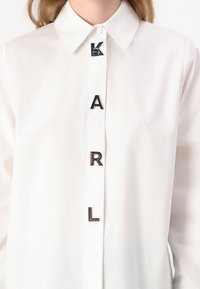 Biała koszula z guzikami, z metalowymi literami układającymi się w napis "KARL" z przodu, z klasycznym kołnierzykiem i długimi rękawami. Gładka tekstura.