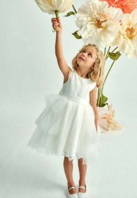 Abito in tulle bianco con un busto texturizzato, gonna a strati e cintura in raso. Bambina che tiene un grande fiore artificiale contro uno sfondo tenue.