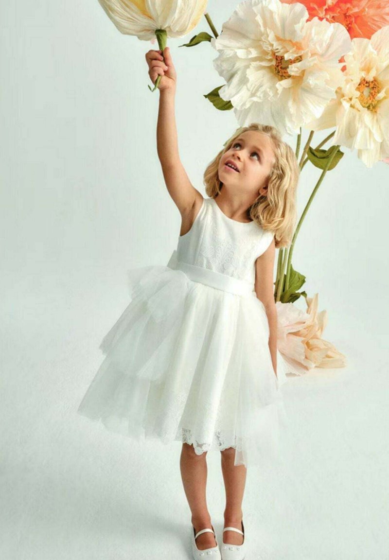 Abito in tulle bianco con un busto texturizzato, gonna a strati e cintura in raso. Bambina che tiene un grande fiore artificiale contro uno sfondo tenue.