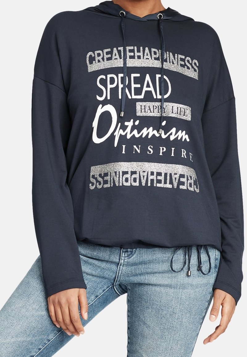 Mörkblå långärmad hoodie med silvertext och grafik. Har en huva, dragsko-stängning och en avslappnad passform. Bärs med ljusblå jeans.