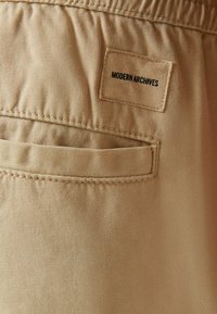 Beige byxor med en slät bomullstextur och ett bakfick. Har en etikett märkta "MODERN ARCHIVES" sydd på midjebandet.