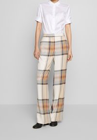 Wijde geruite broek in crème, met oranje en marineblauw ruitpatroon. Gecombineerd met een aansluitend witte overhemd met knopen.