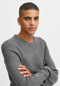 Pulover gri din tricot cu un model texturat, decolteu rotund și manșete elastice. Modelul are brațele încrucișate, evidențiind croiala și designul puloverului.